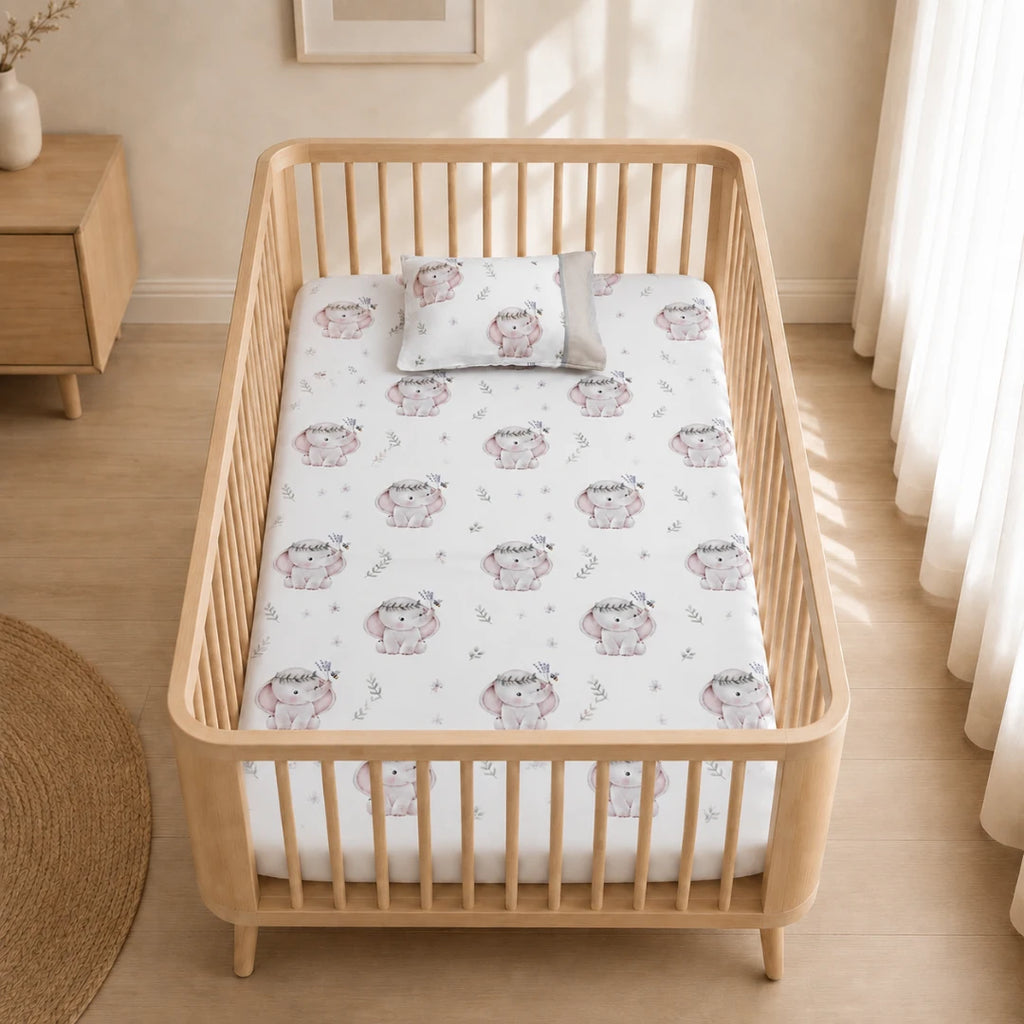 Sábana ajustable para cuna de bebé con funda de almohada, estampado de elefantes rosa y detalles florales sobre fondo blanco, colocada en una cuna de madera . Juego de ropa de cama infantil de polialgodon suave, ideal para colchón de cuna y habitación de bebé.